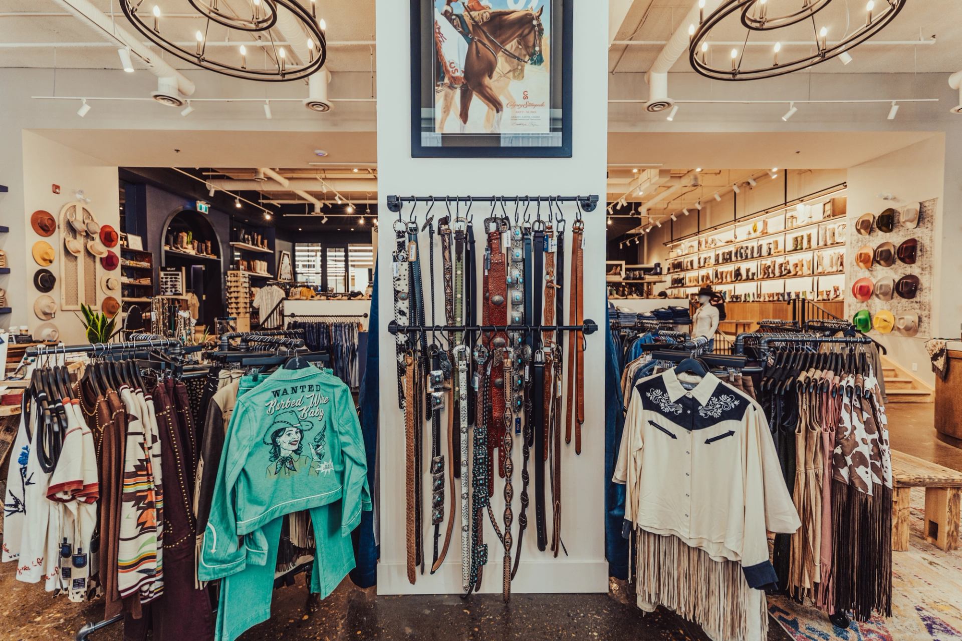 Western clothing store interior with belts, fringed shirts, hats and jackets on display racks.