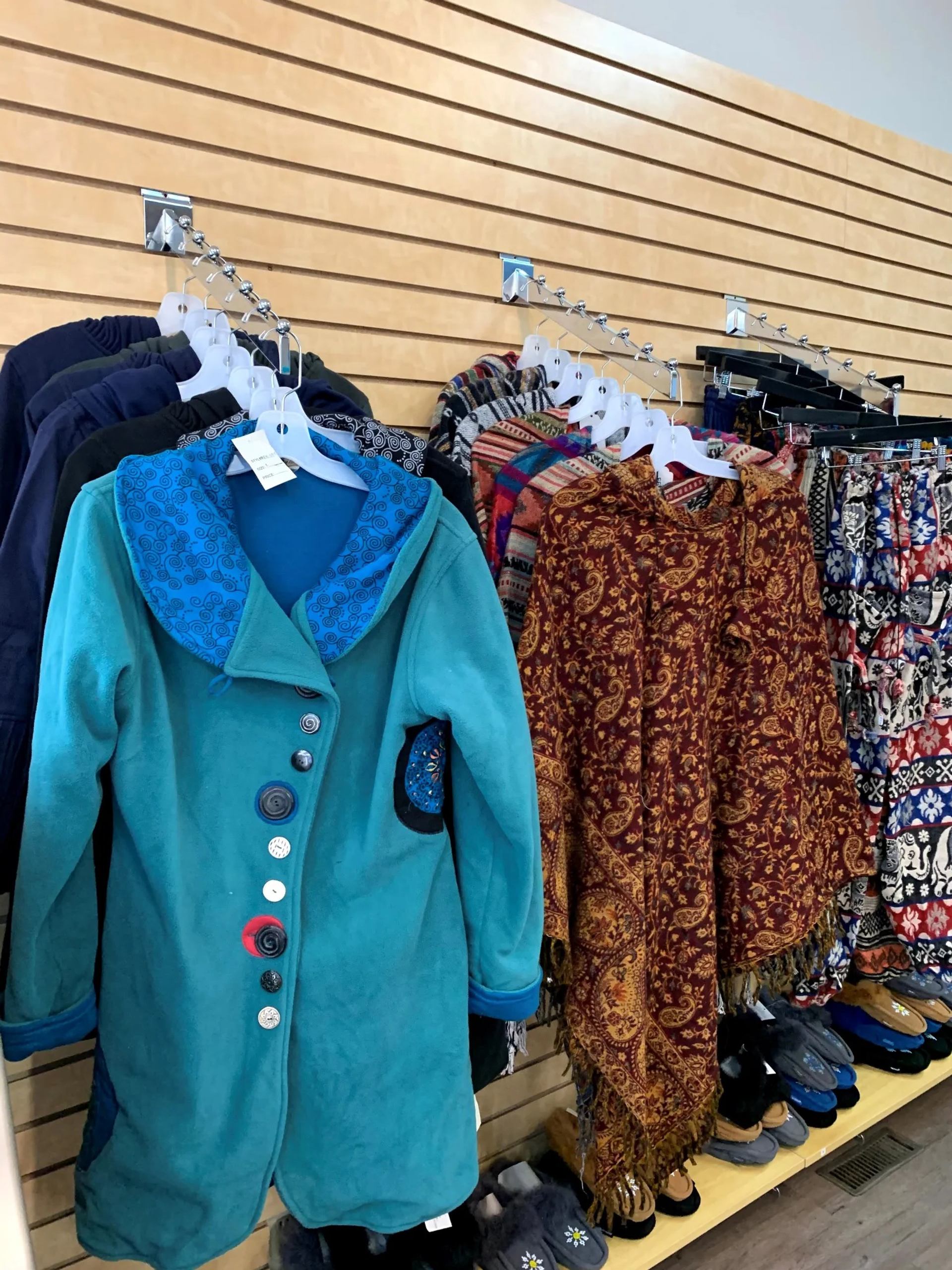 Clothing rack with teal coat, patterned shawls and slippers in a retail shop.