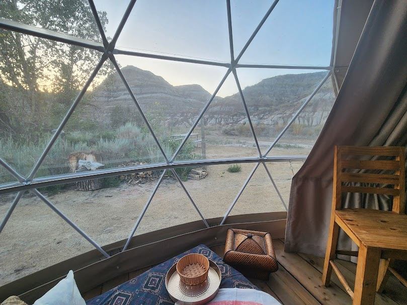 Geodesic dome interior with bed and chair overlooking dry hills and sparse vegetation.
