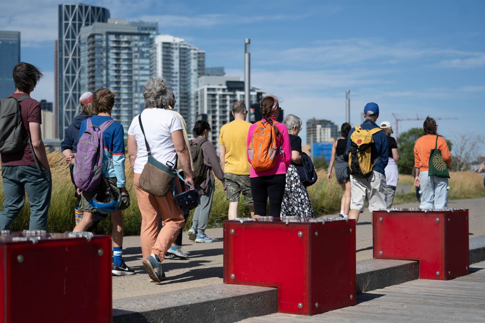 People with backpacks walk along a path beside red benches and tall buildings.