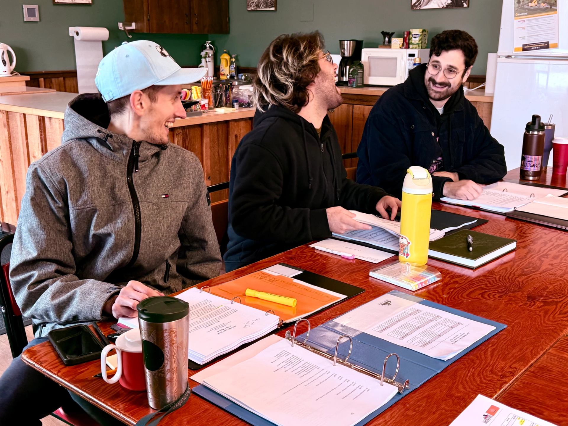 Three people discuss documents in binders around a wooden table.
