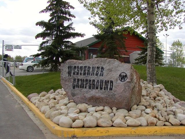 Stone sign reading “Westerner Campground” beside trees, red building and parked cars.