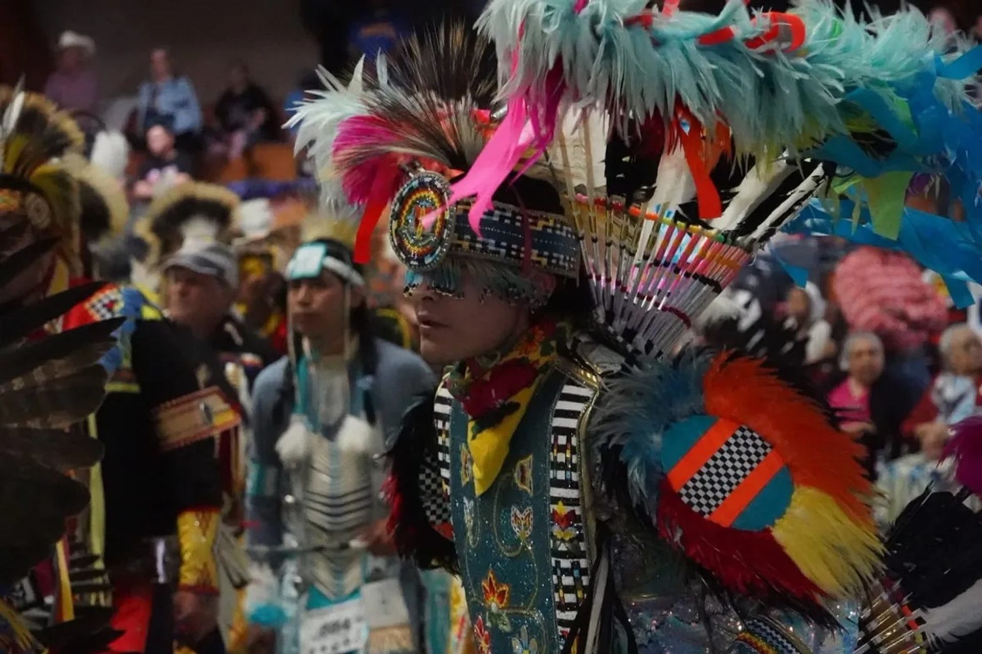 Powwow dancer in colourful feathered regalia performs among other participants indoors.