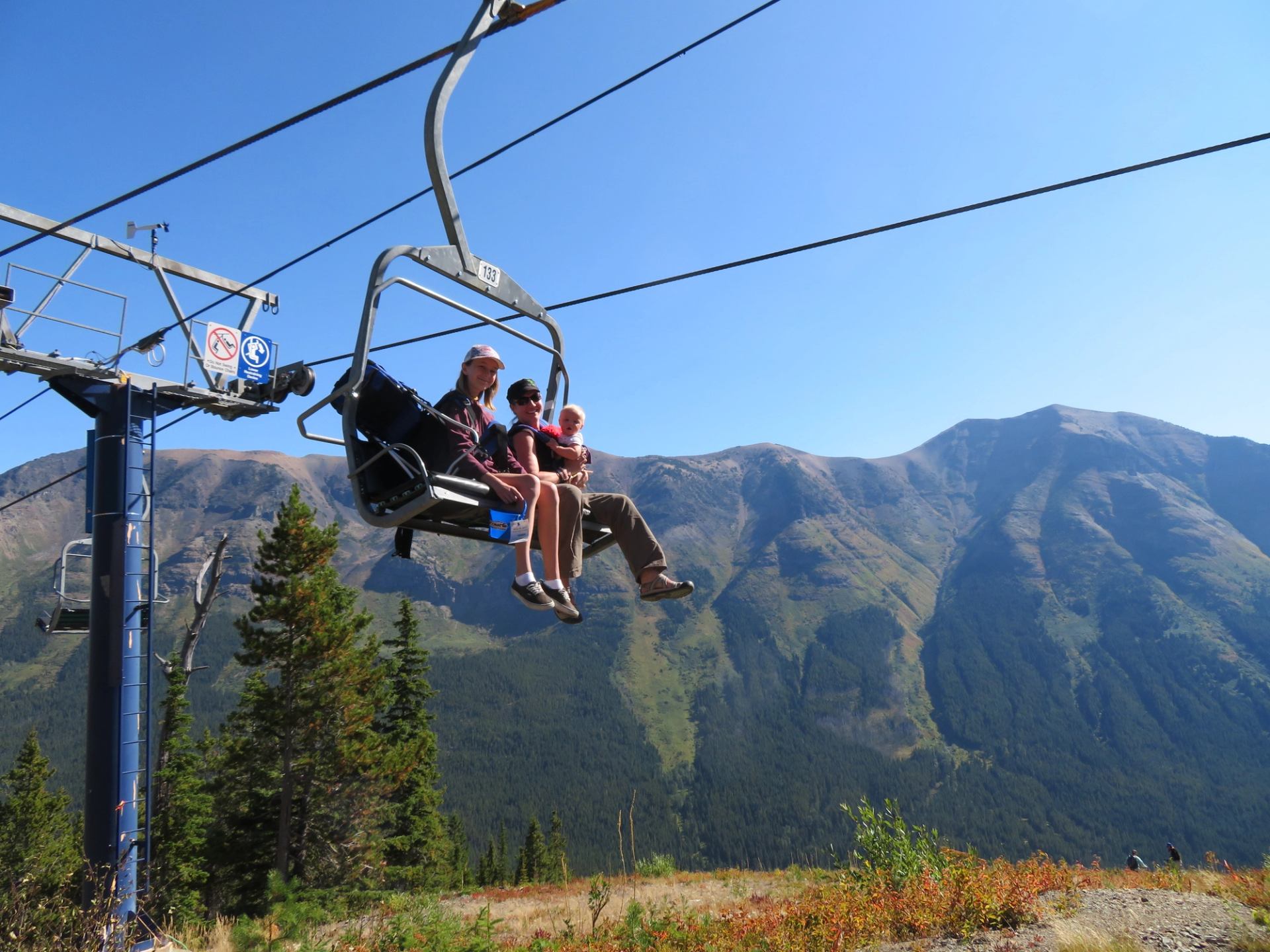 Chairlift carries three people above forested mountain slope under clear blue sky.