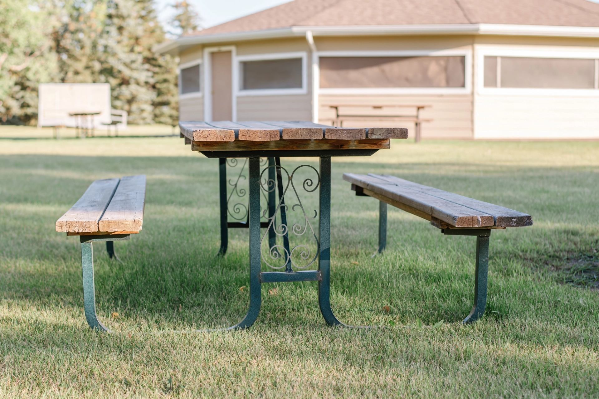 Wooden picnic table with benches on grass, with a light-coloured building behind.