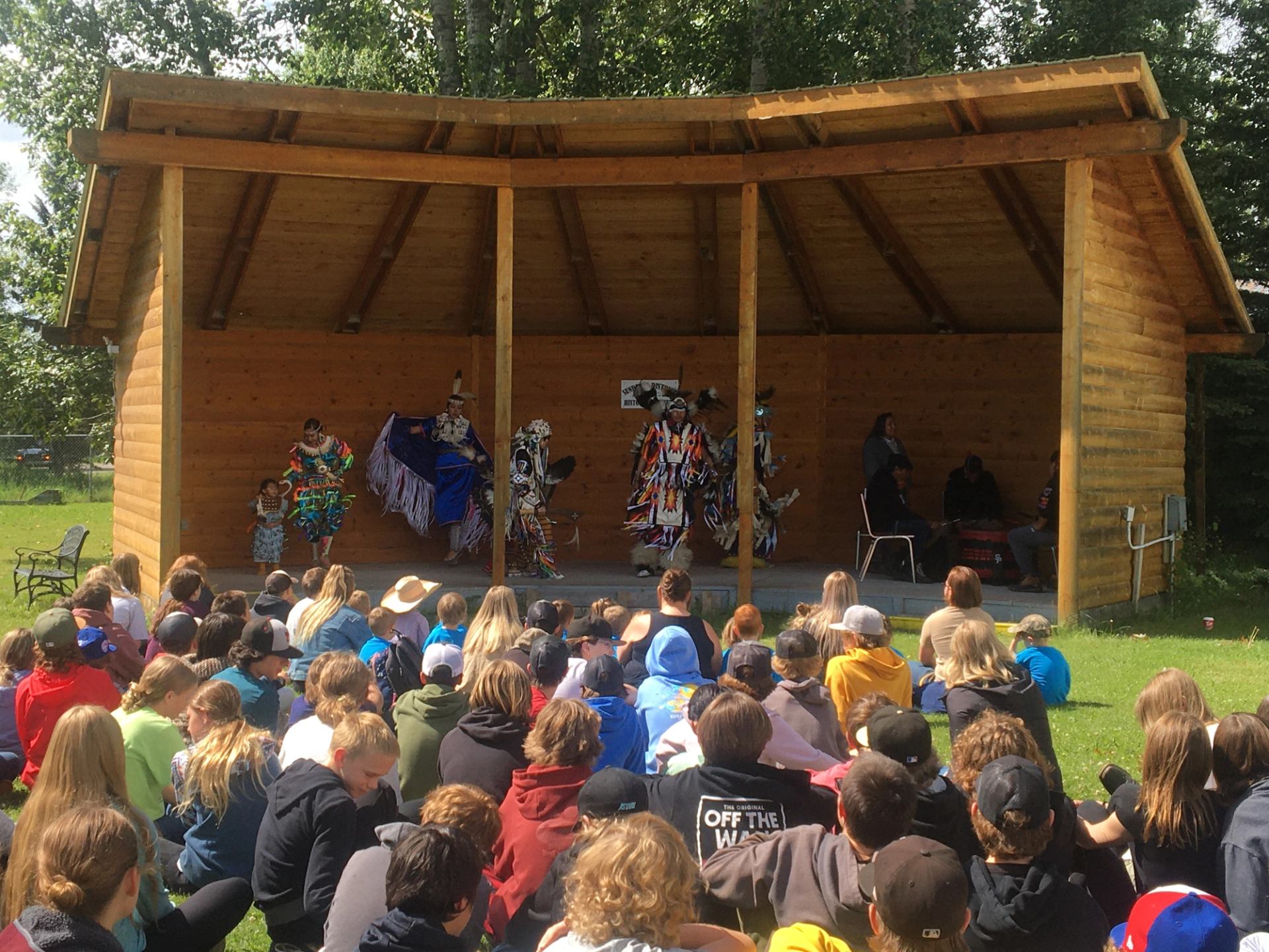 An audience of youth sits on the grass, watching Indigenous dancers perform in vibrant regalia on an outdoor wooden stage. A group of drummers and singers accompany the performance from the side of the stage.