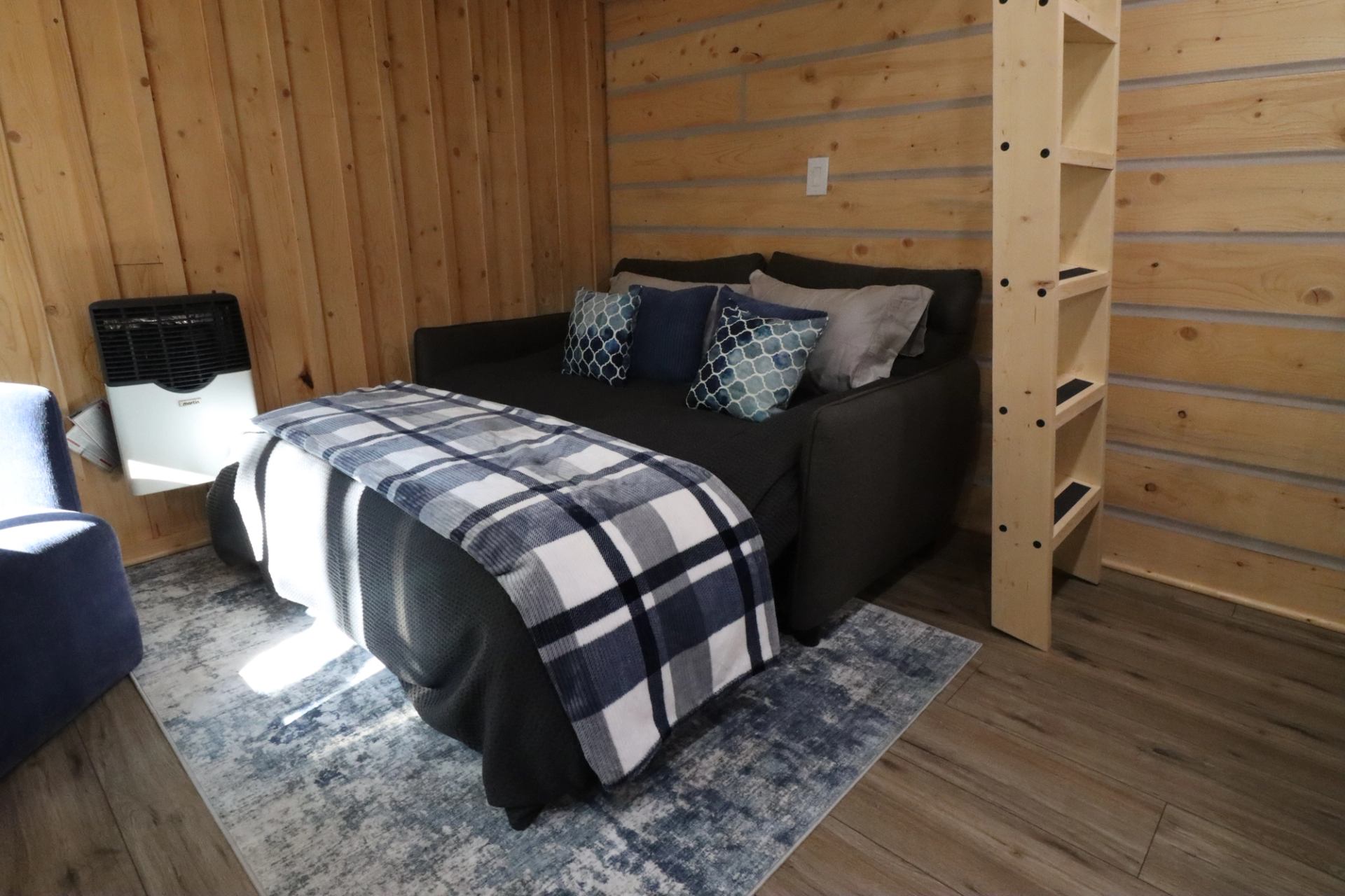Cozy room with sofa bed, pillows, blanket, wall heater, empty bookshelf, and blue rug.