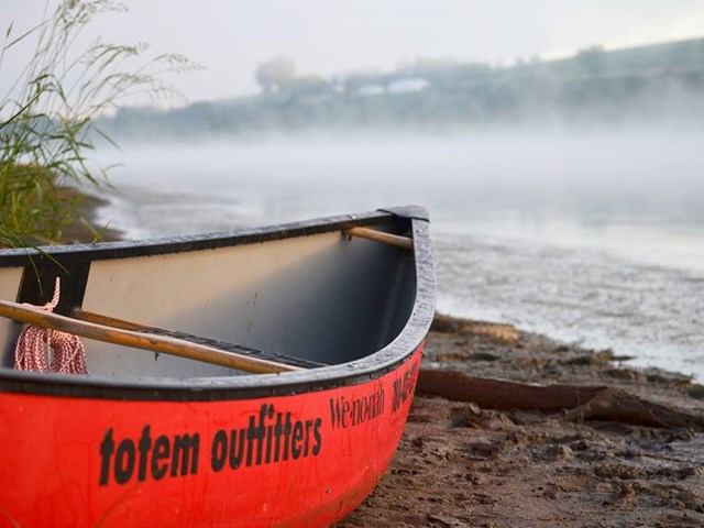 Red Totem canoe on misty lakeshore with plants and blurred trees in background.