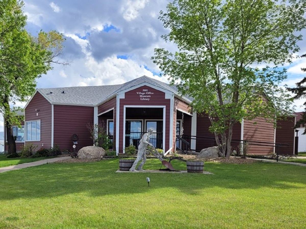 A red building with the sign "Village Office, Museum & Library" on it. A statue of a person working is in front of the building, surrounded by trees and a grassy area.