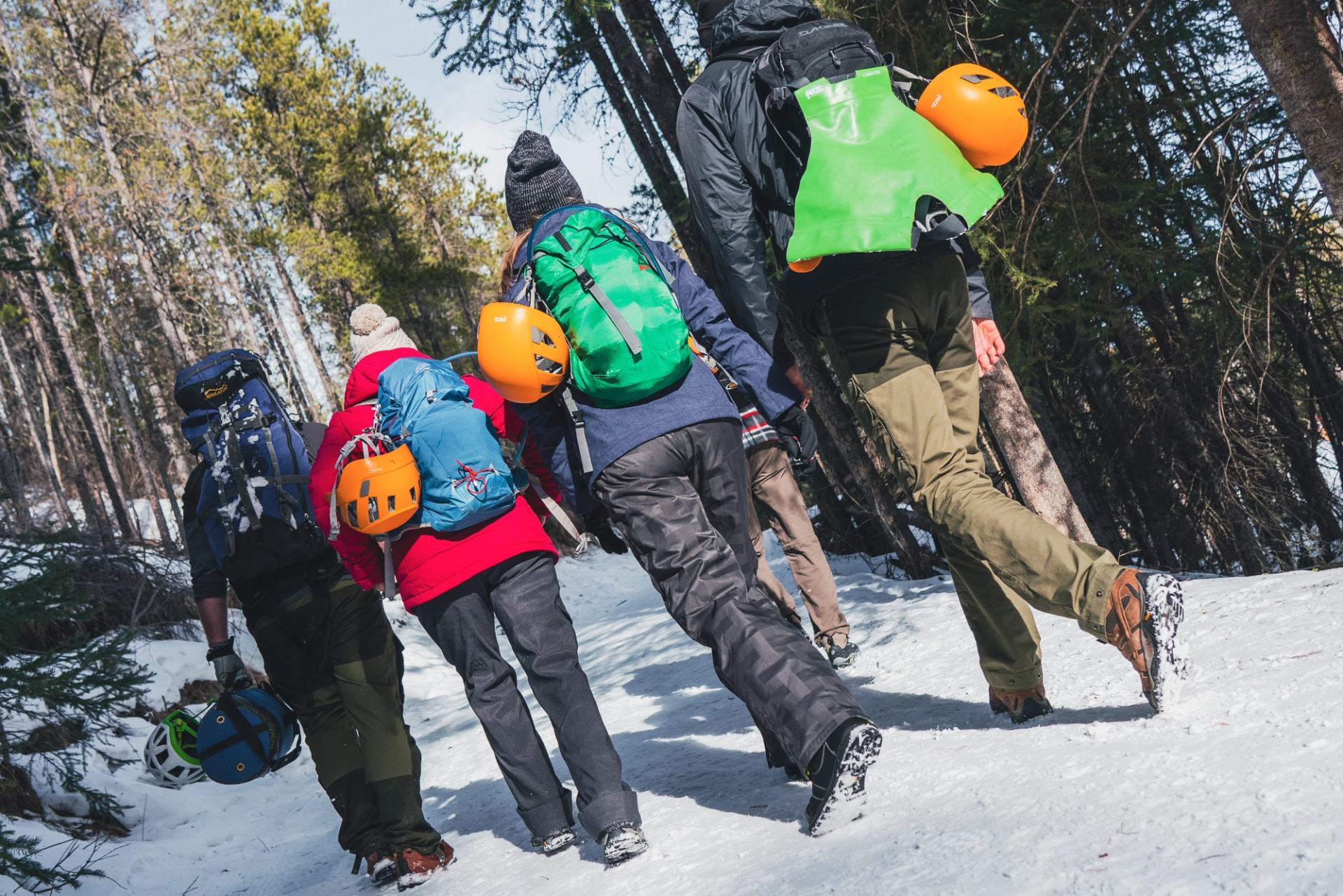 Participants walking uphill on a snowy forest trail carrying helmets and gear.