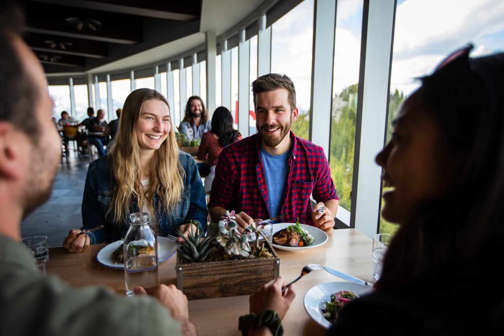 Group of people eating at a restaurant table beside large windows overlooking trees.