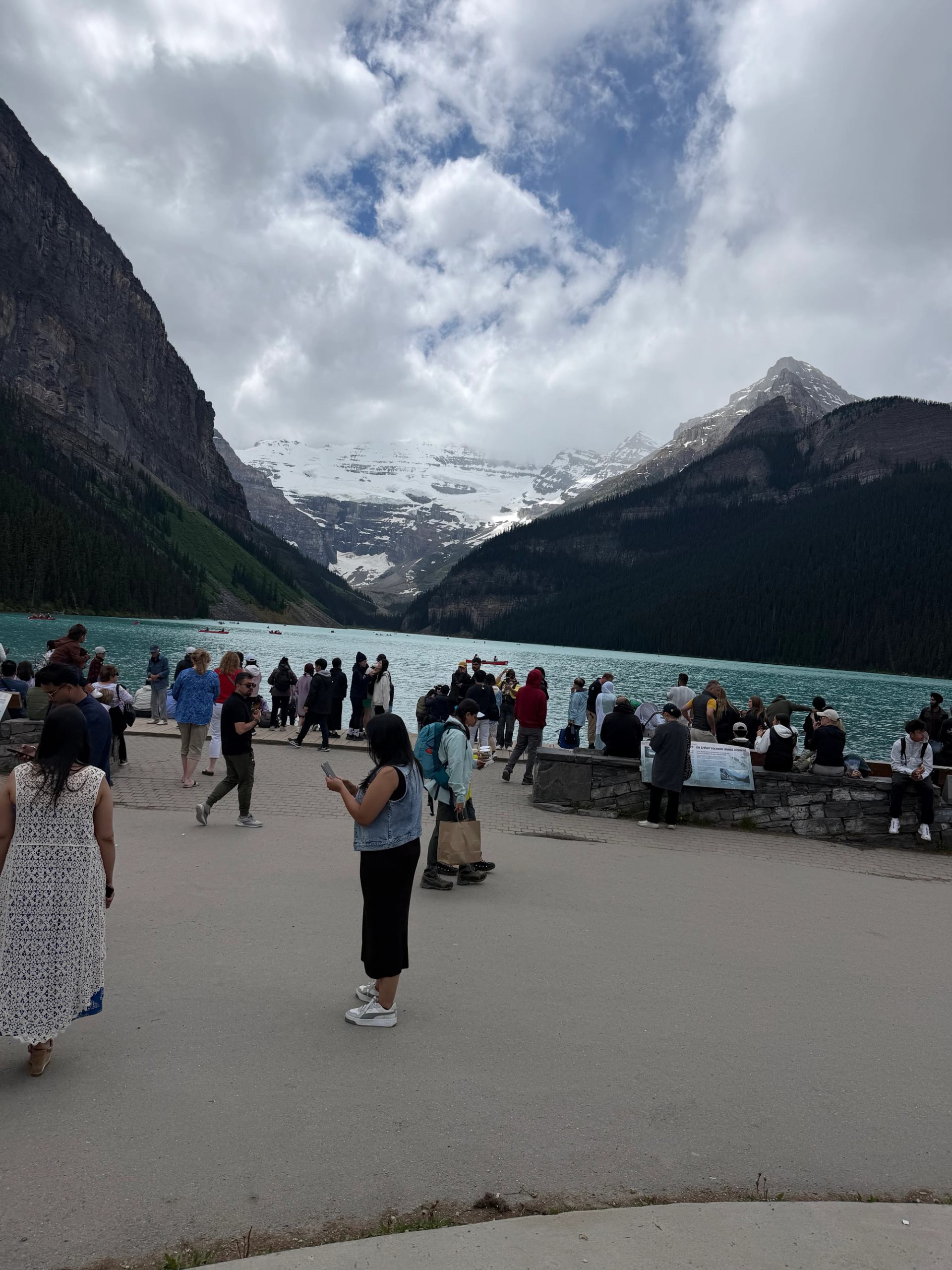 Tourists gather by turquoise mountain lake with snow-capped peaks and cloudy sky.