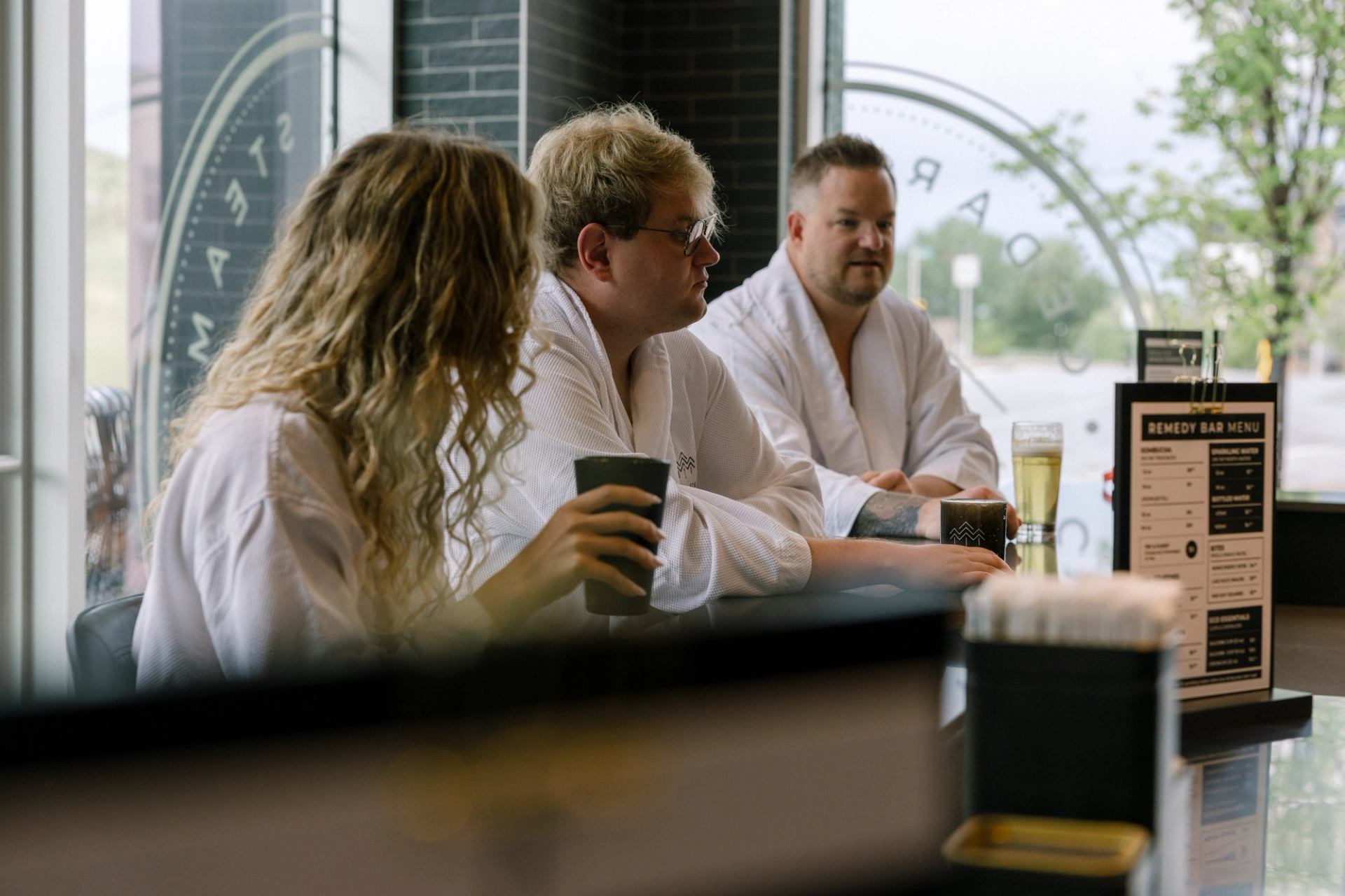 People in white robes sit at a bar, drinking beverages beside a menu.