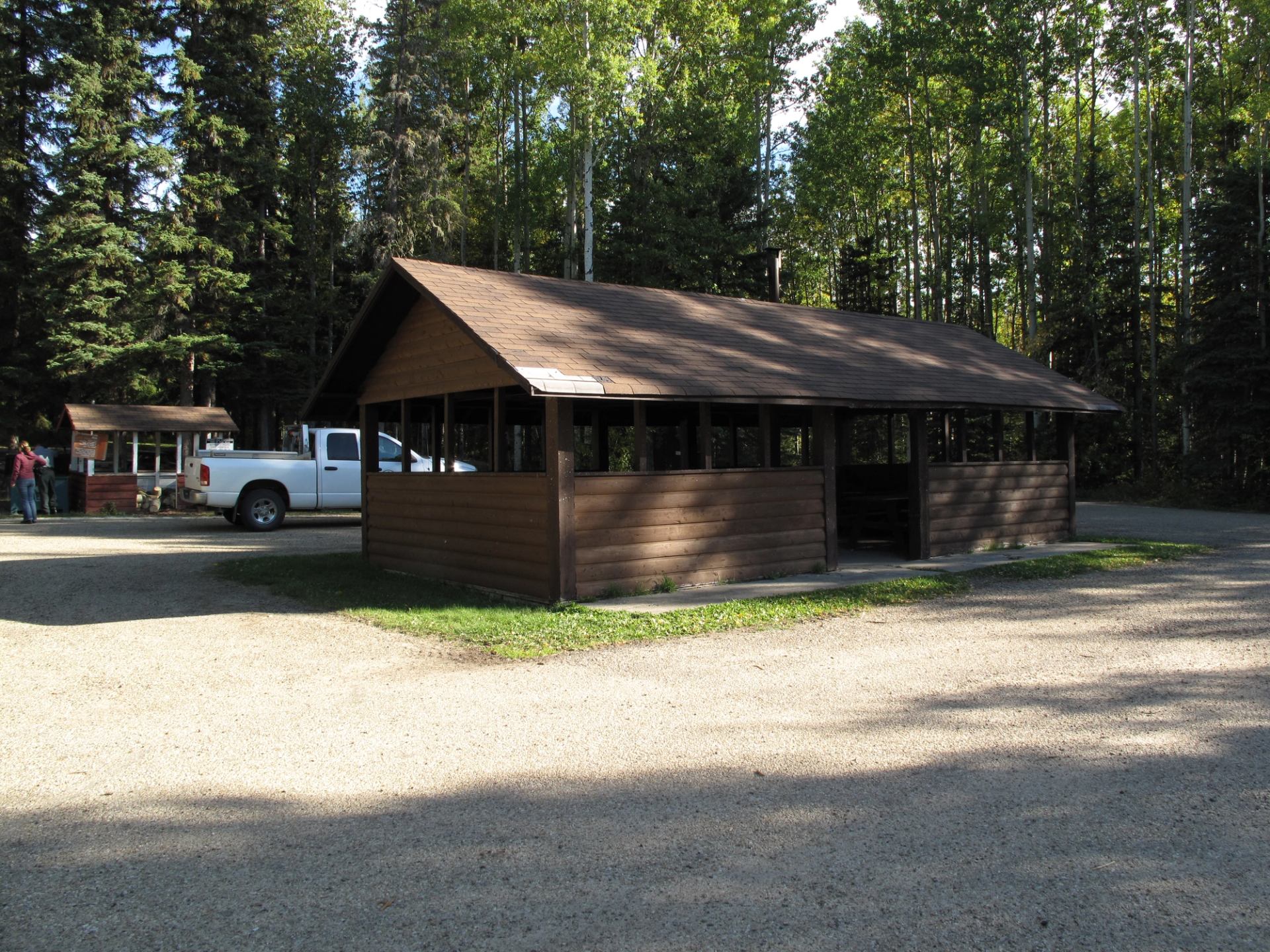 Wooden shelter building in a forested campground with vehicles nearby.