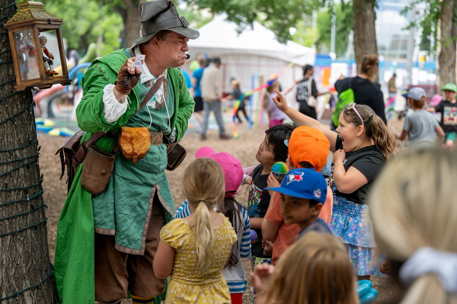 Performer in green costume entertains children outdoors at a crowded festival area.