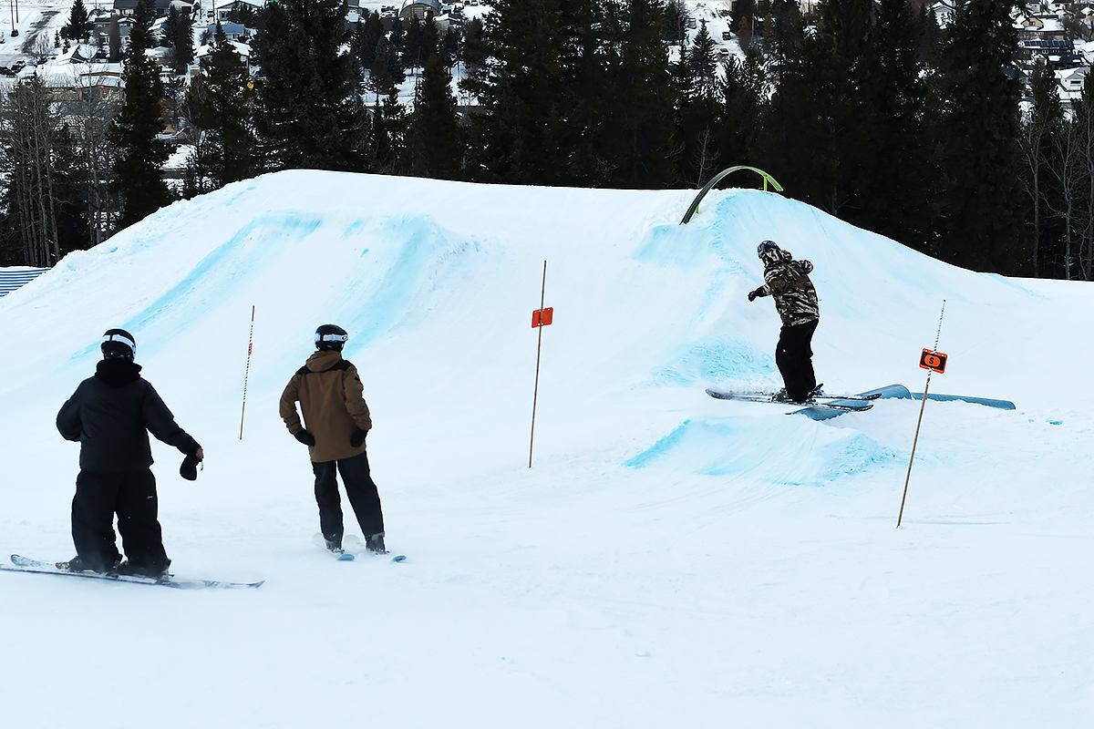 Snowboarders riding through a terrain park as one person hits a small feature.