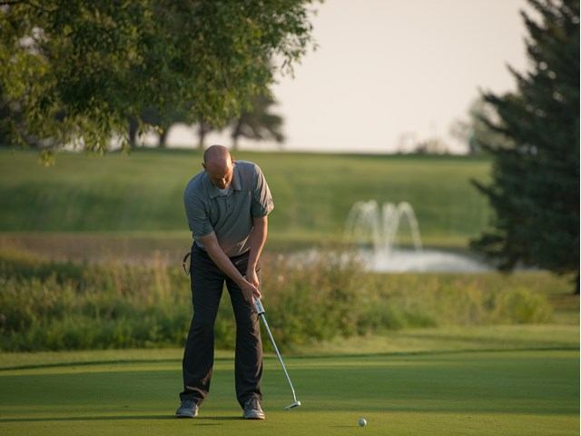Golfer putting on a green near a fountain, with trees in the background.