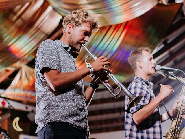 Musicians play trumpet and trombone onstage at a festival with colorful backdrop and banners.