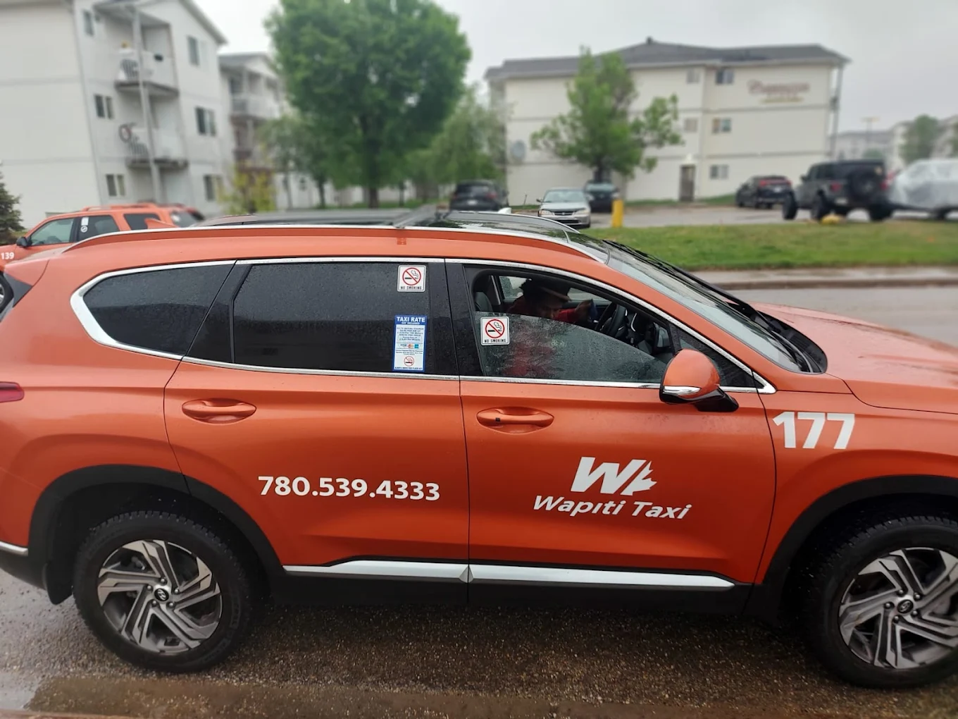 Orange Wapiti Taxi SUV with a driver on a wet residential street.