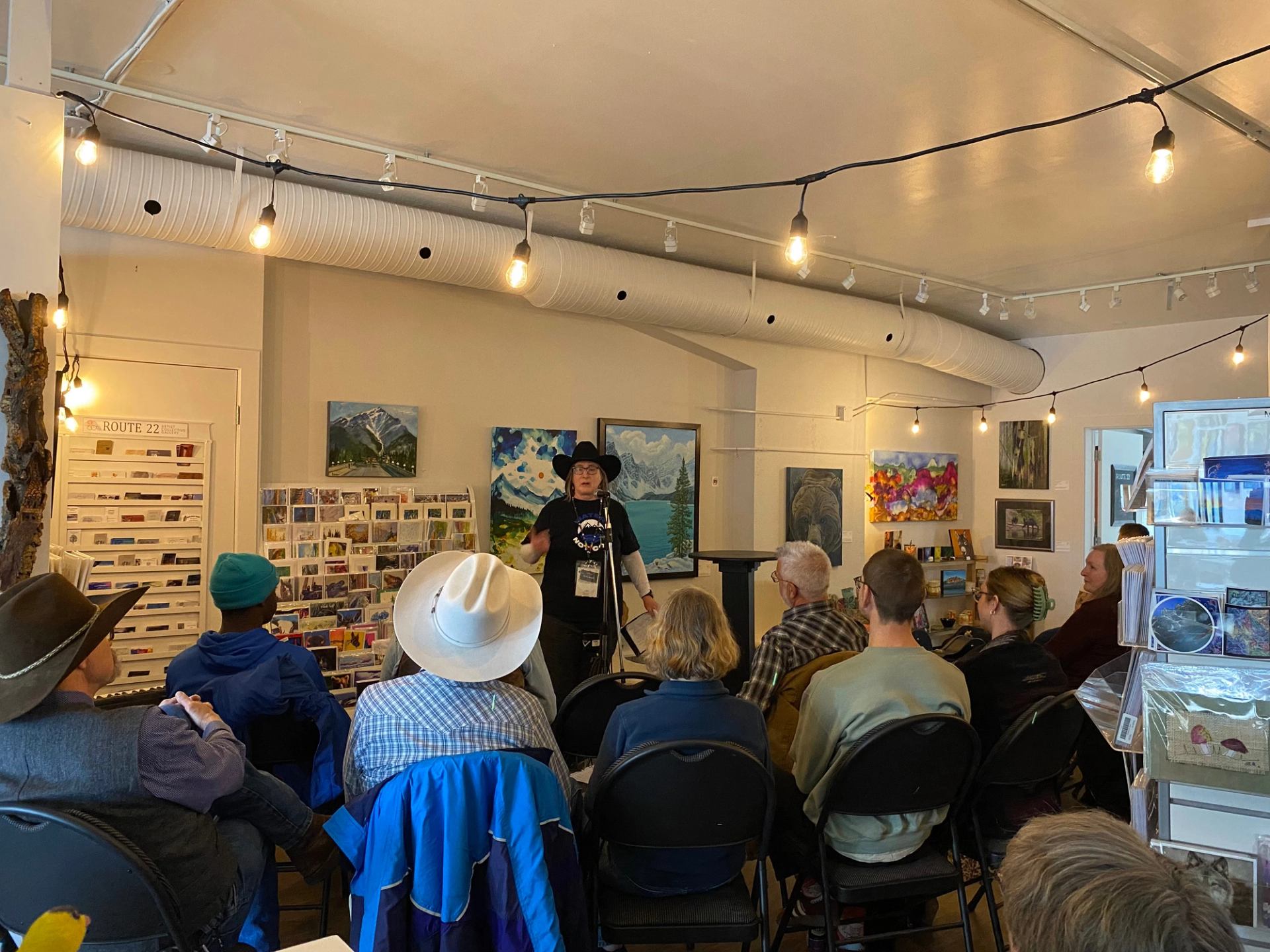 Speaker addresses a seated audience in an art gallery under string lights.