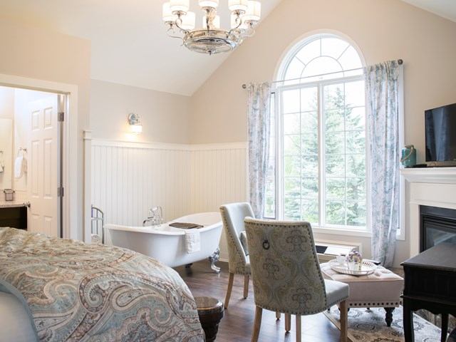 Elegant bedroom with fireplace, chandelier, and freestanding tub near arched window.
