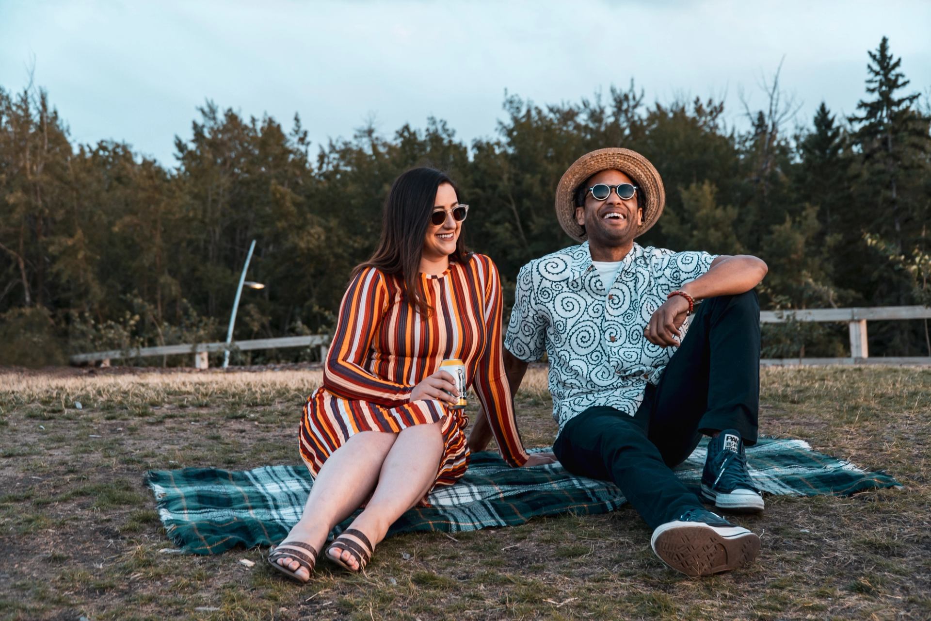 A smiling couple sits on a picnic blanket outdoors, with a man in a hat and a woman holding a drink.
