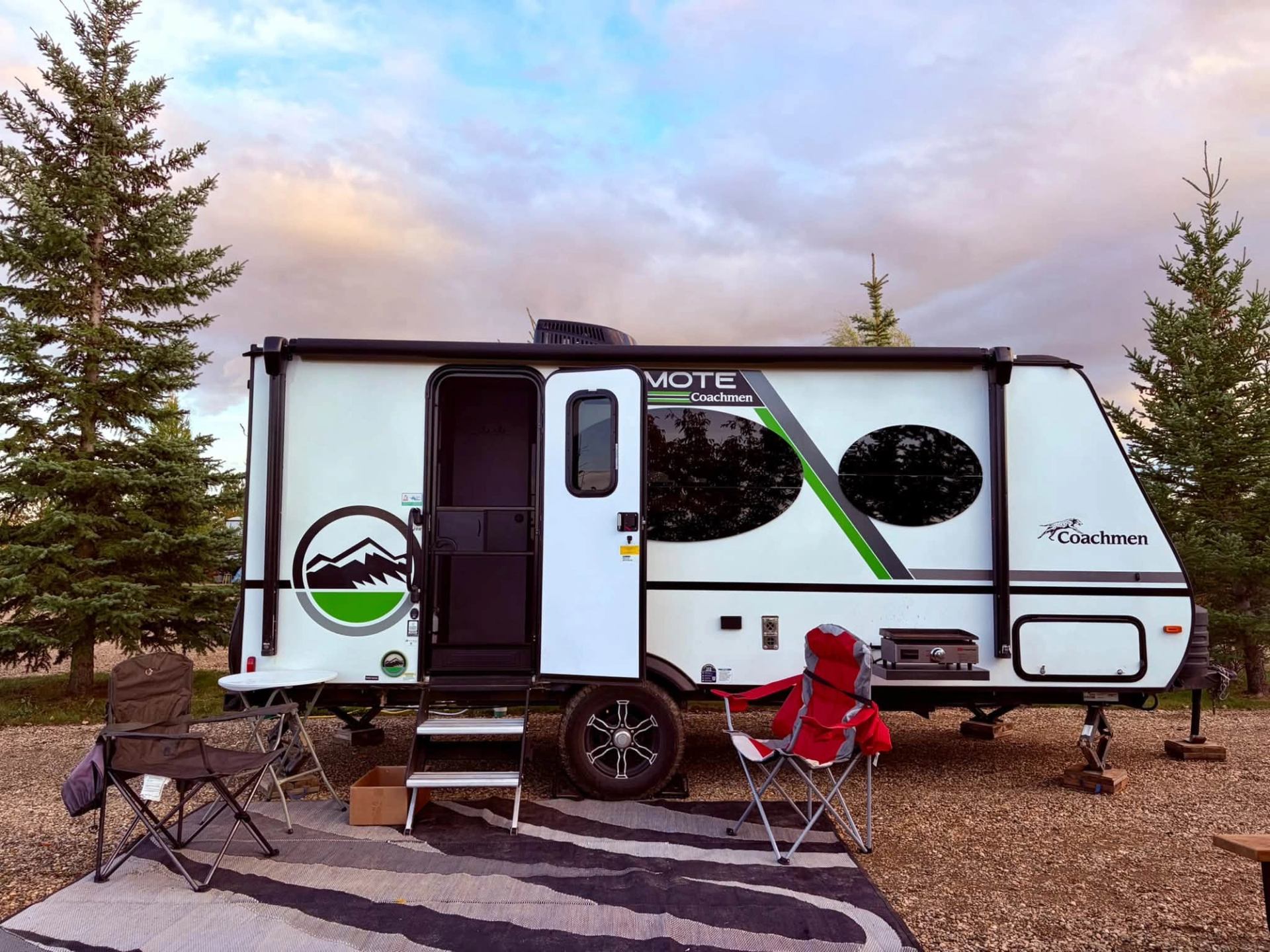Coachmen travel trailer with camping chairs, table and rug among evergreen trees.