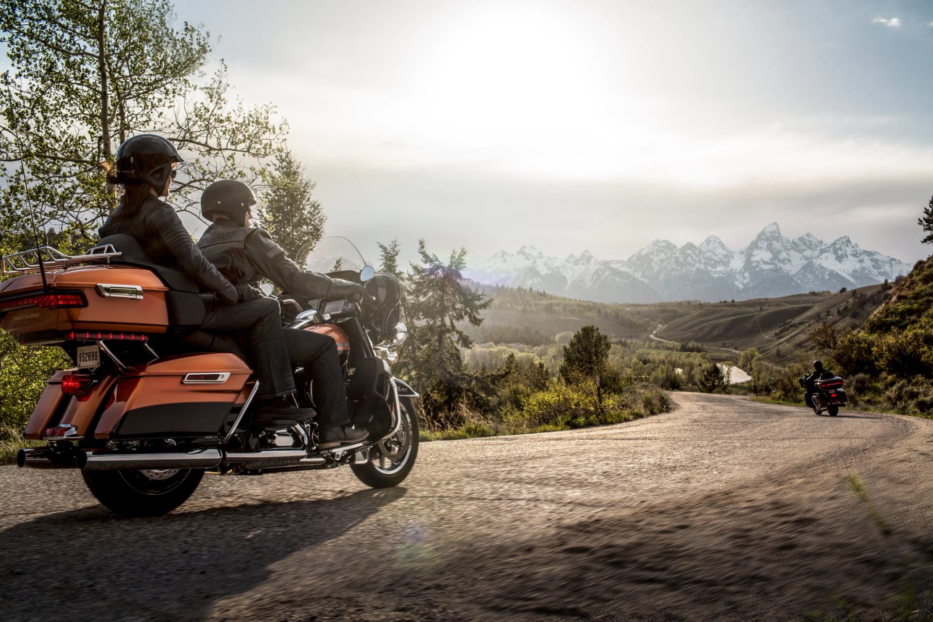 Motorcyclists ride along a winding rural road toward snow-capped mountains.