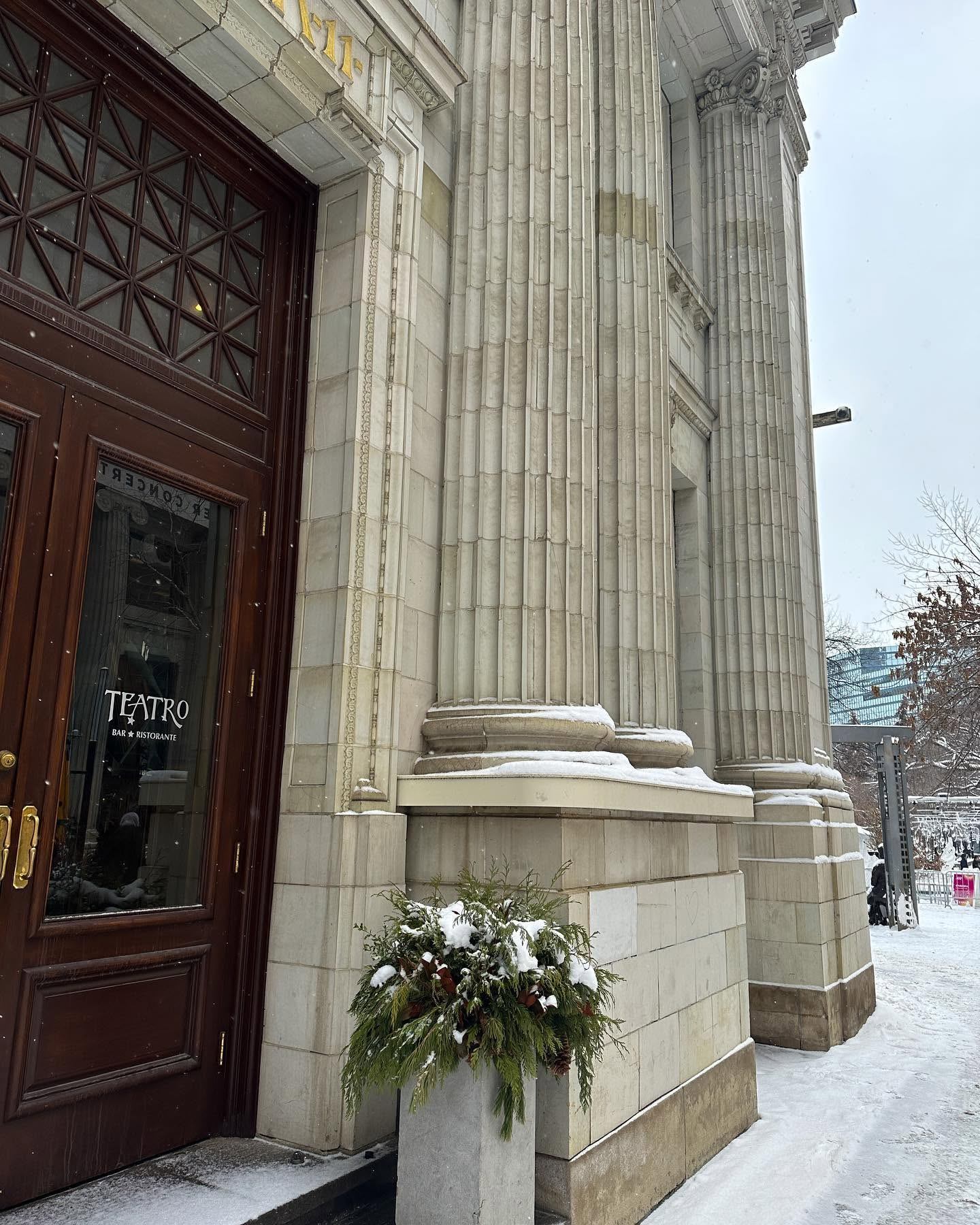 Stone building entrance with Teatro door, tall columns and snowy planter outside.