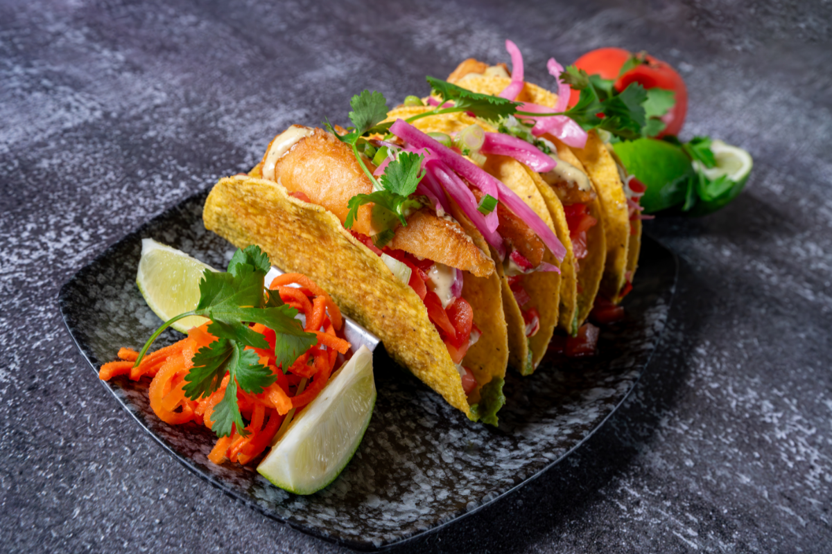 Grilled tacos with pickled onions, cilantro, and lime wedges on a dark plate.