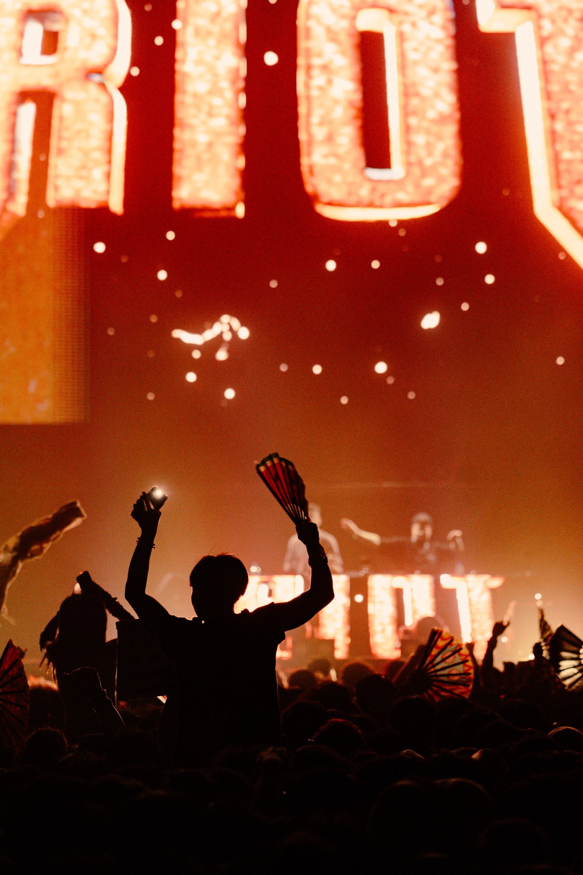 Silhouettes of concertgoers waving fans and raising hands in front of a glowing stage.