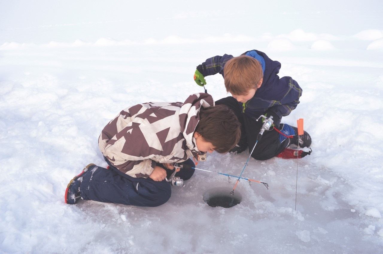 Children kneel on snowy ice, holding fishing rods over a round ice fishing hole.