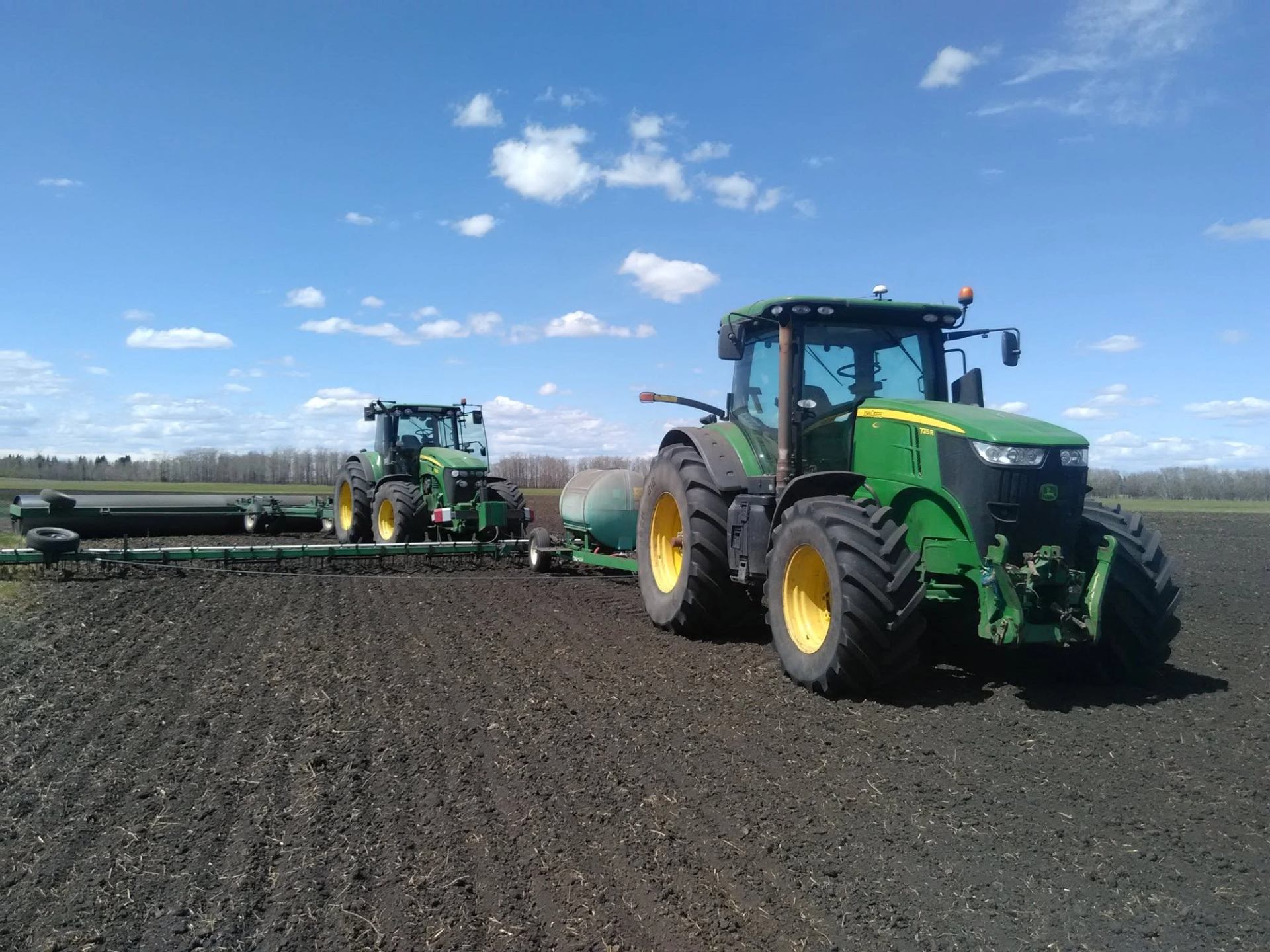 Two large tractors work across a freshly prepared field under a bright blue sky with scattered cloud