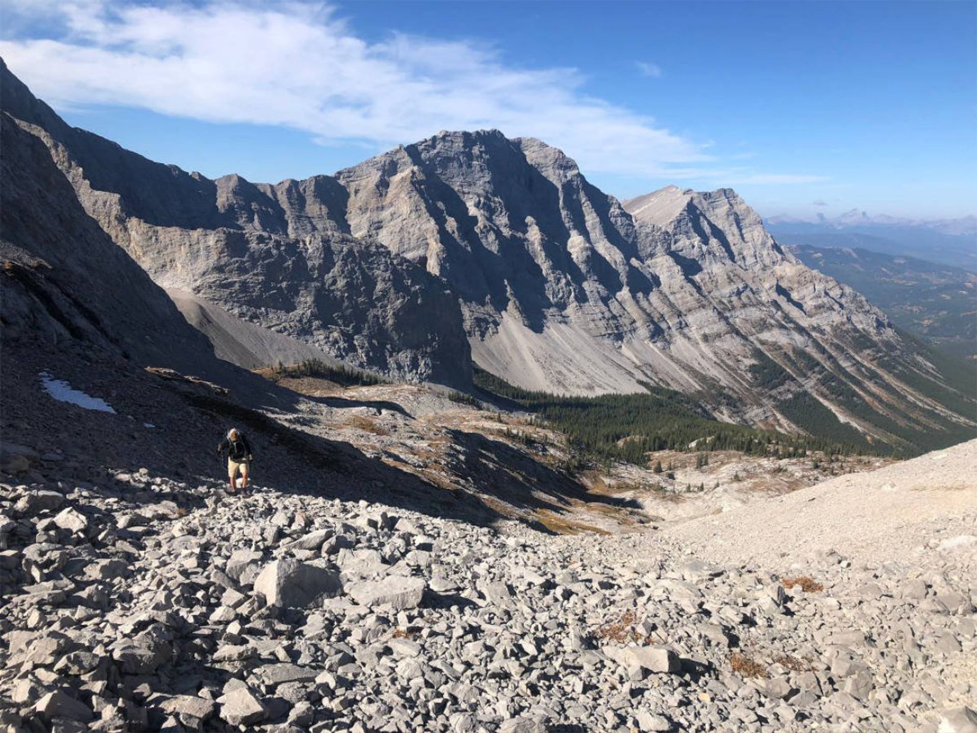 Hiker climbs rocky mountain slope with jagged grey peaks and distant forested valley.