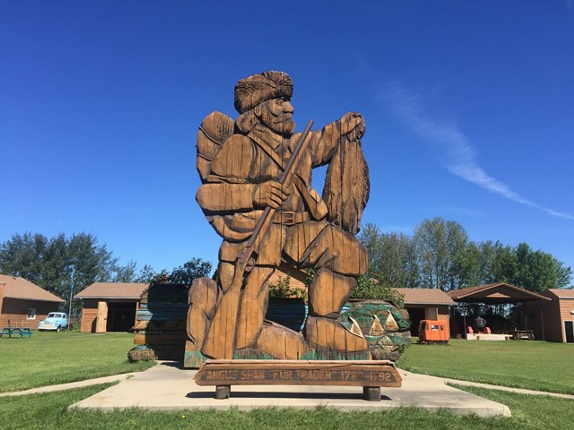 A large wooden statue of a frontier man holding a rifle and animal pelt against a clear blue sky. Surrounded by green grass and buildings.