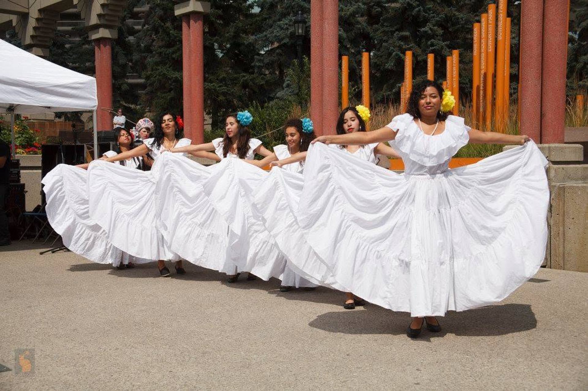 Dancers in white traditional dresses hold wide skirts during outdoor performance by columns.