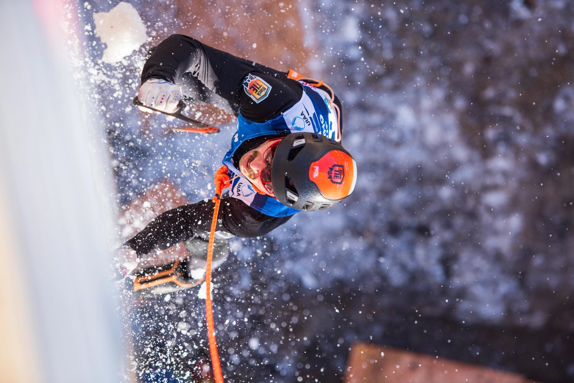 Ice climber scaling frozen wall with gear in snowy competition setting.