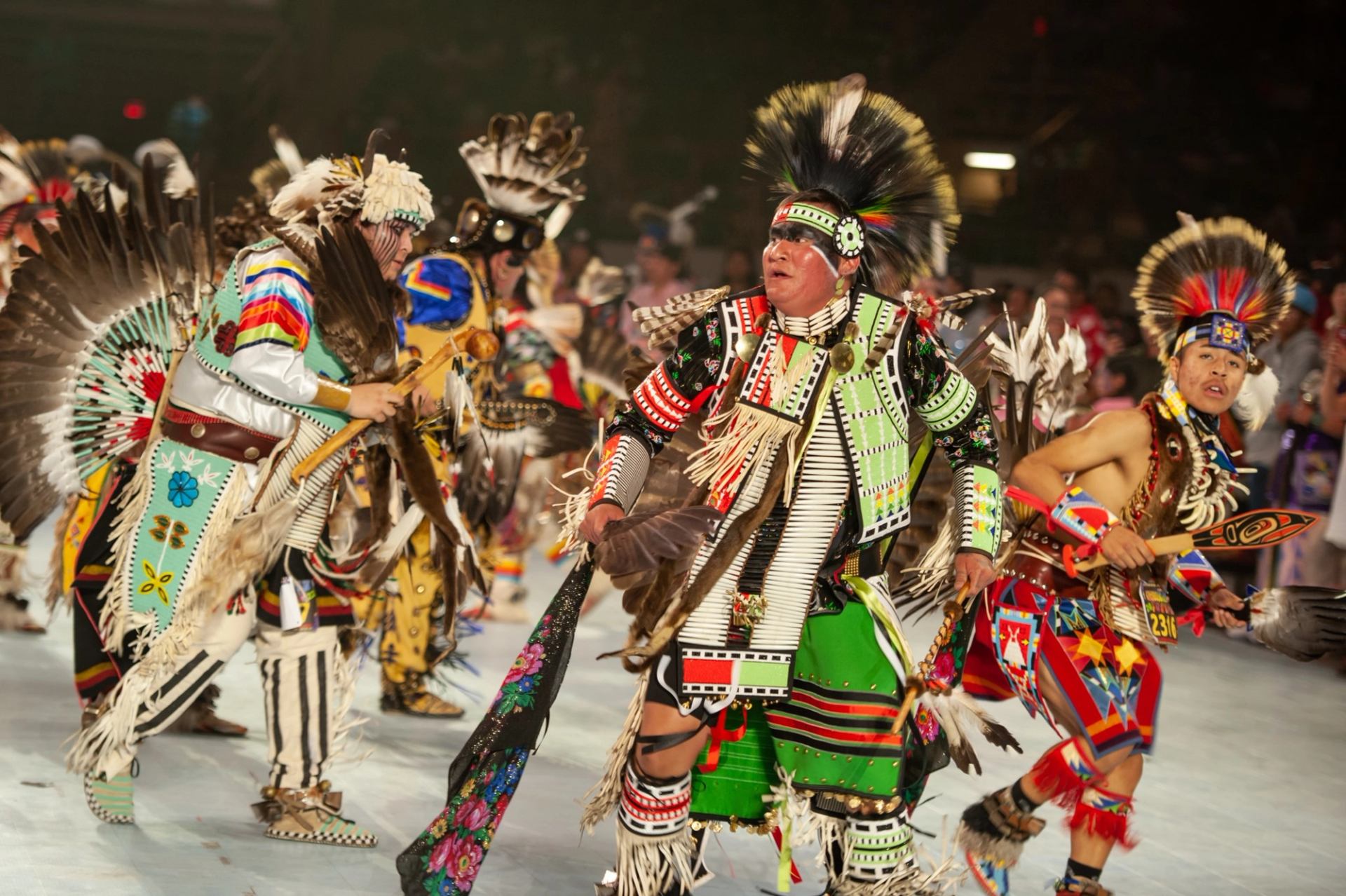 Indigenous dancers in ornate regalia perform during a powwow, with drum and crowd behind them.