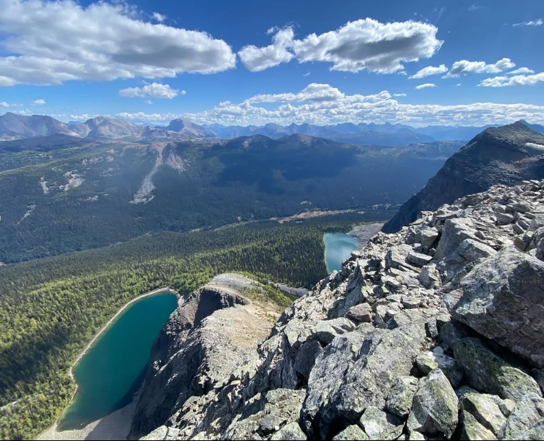 Rocky mountain ridge overlooks turquoise lakes, forested valleys and distant peaks.