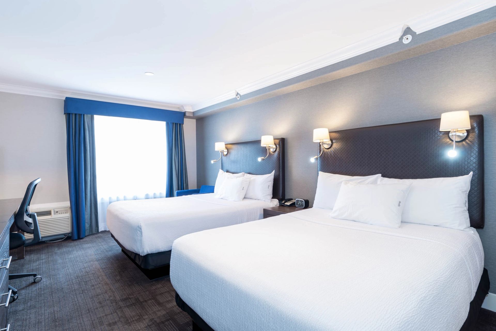 Guest room with two beds, bedside lighting, work desk, and large window.
