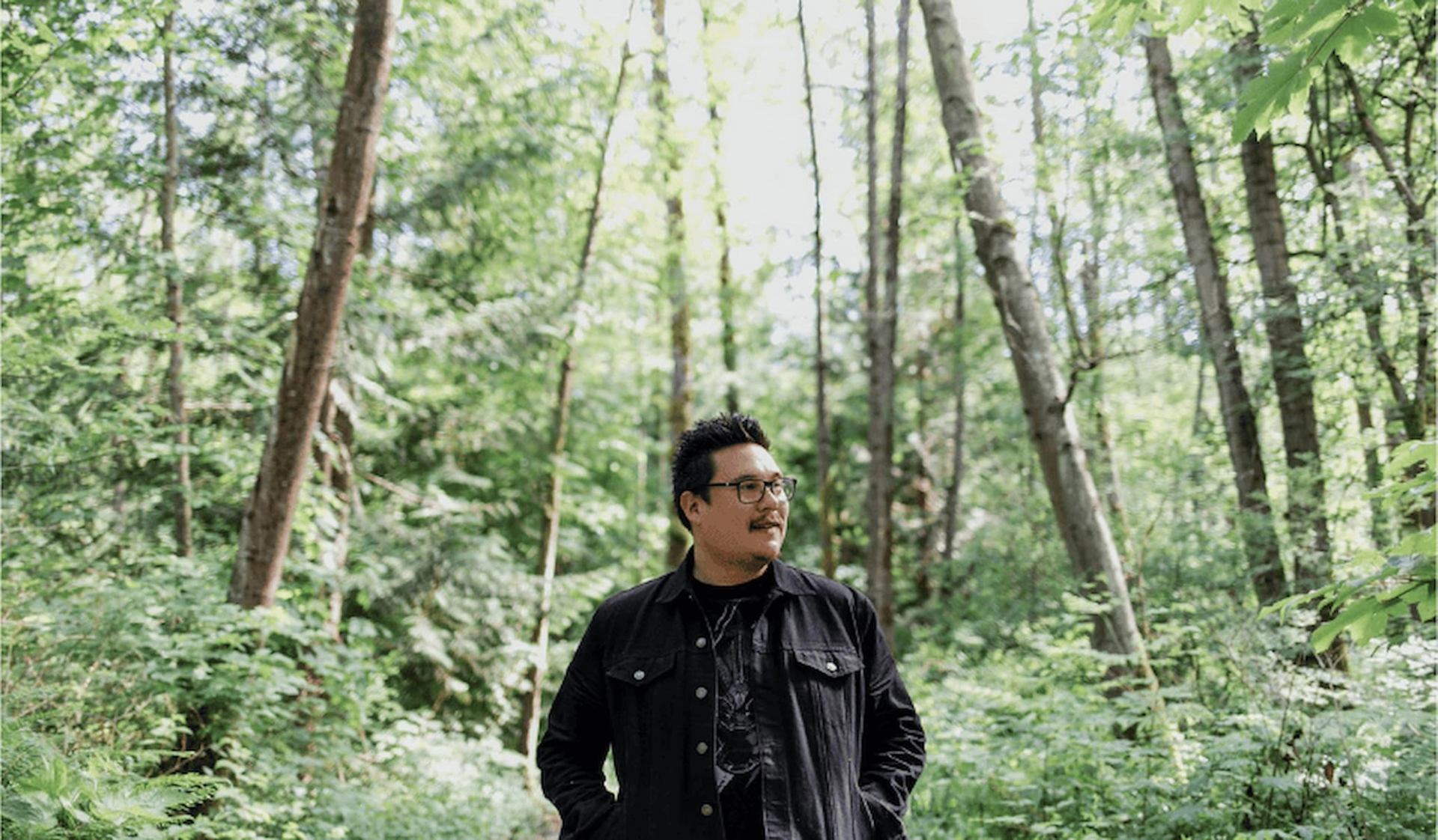 Person wearing glasses and dark jacket standing in a dense green forest.