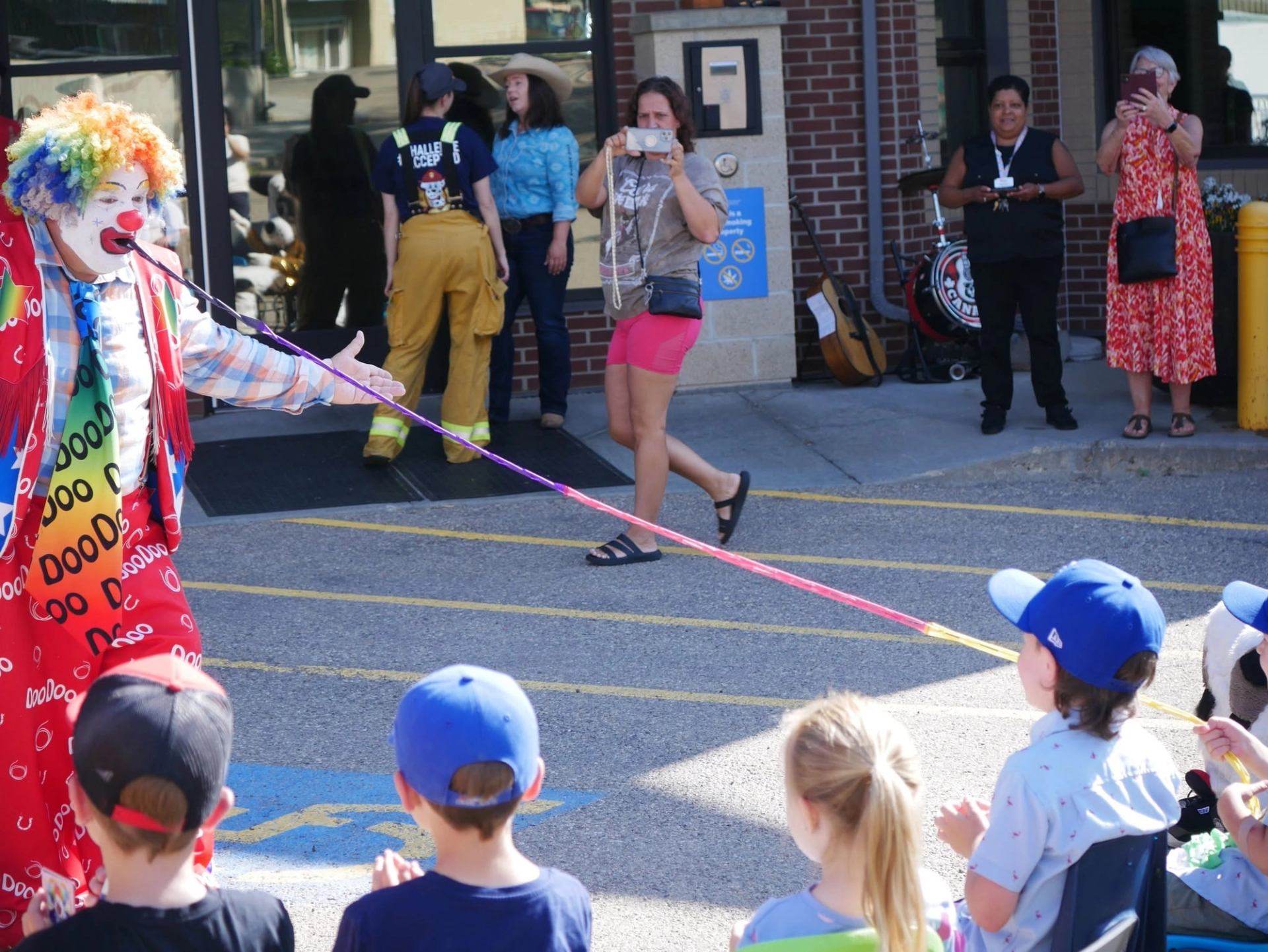 Colourful clown performs with a ribbon as children and adults watch.