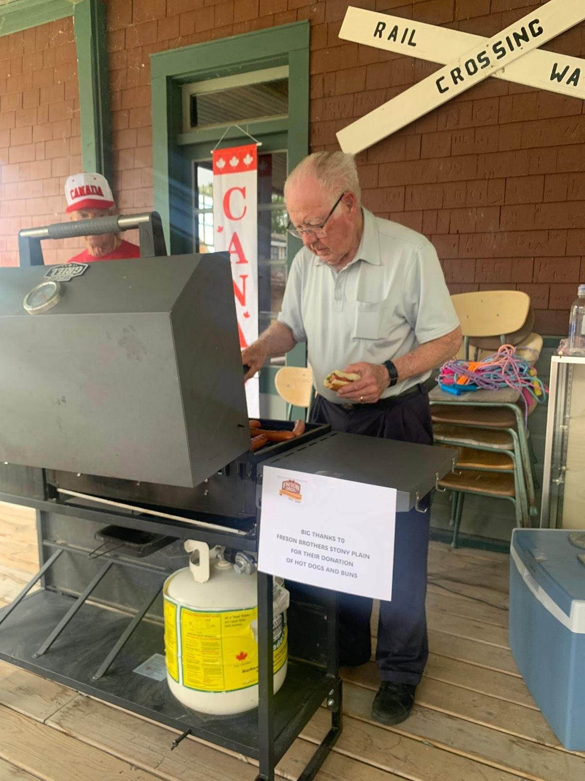Man grilling hot dogs at outdoor event with Canada sign and rail crossing decor.