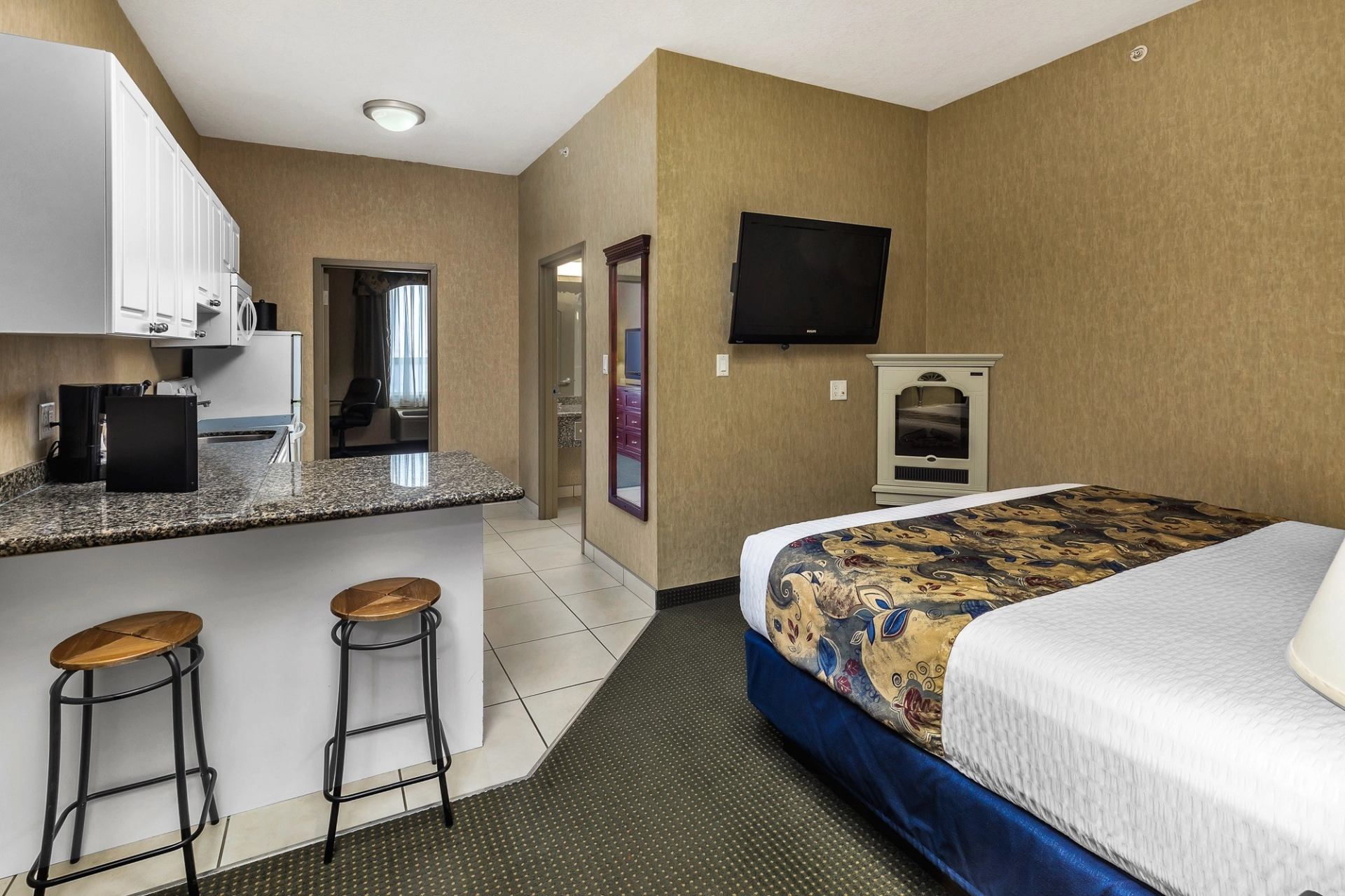 Hotel room kitchenette with bar stools, bed, wall TV, and coffee table near doorway.