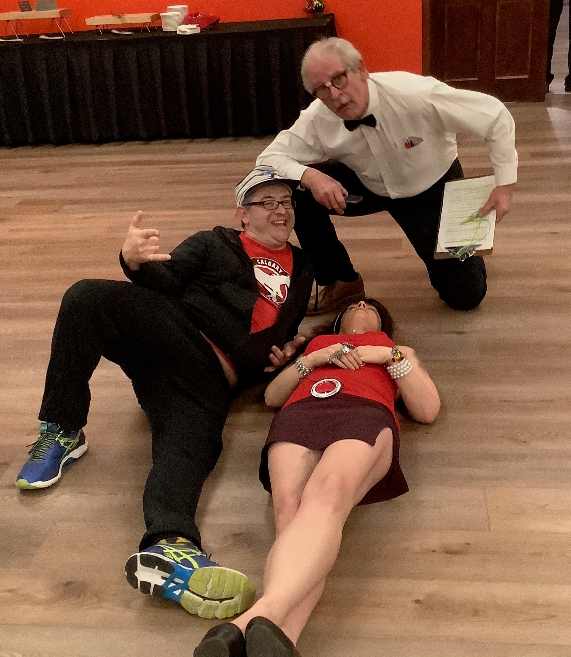 Three people pose on a wooden floor, with two reclining and one kneeling.