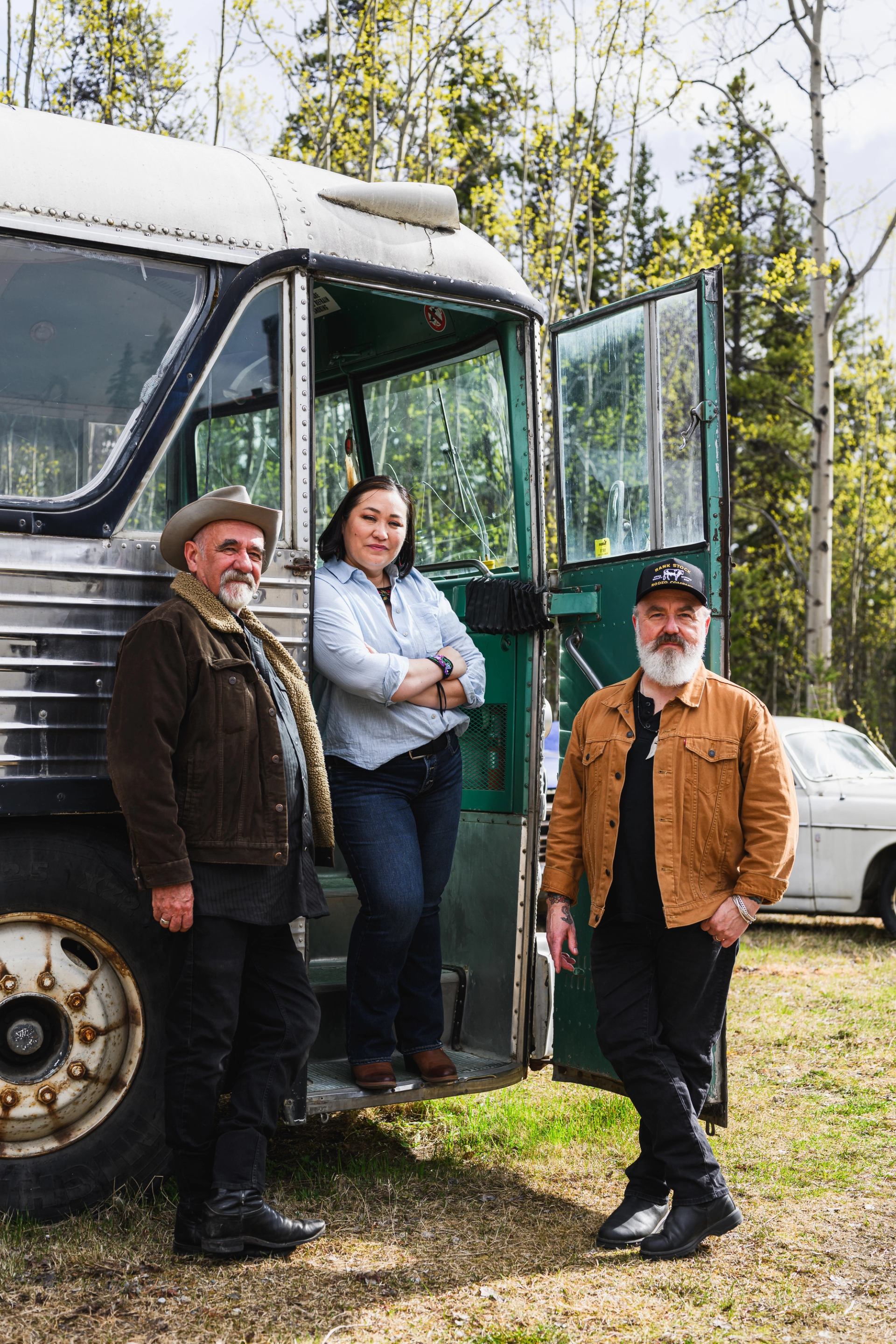 Three people stand by an old bus in a forested outdoor area.