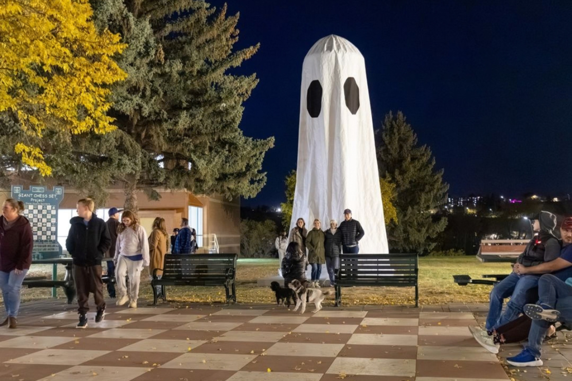 People and dogs gather near a towering white ghost decoration in a park.
