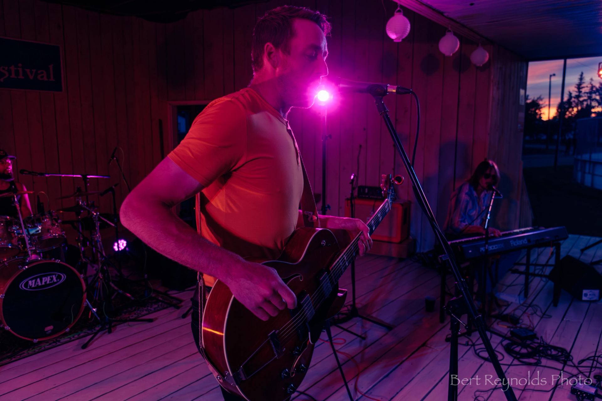 Guitarist sings into microphone under purple stage lights with drummer and keyboardist behind.