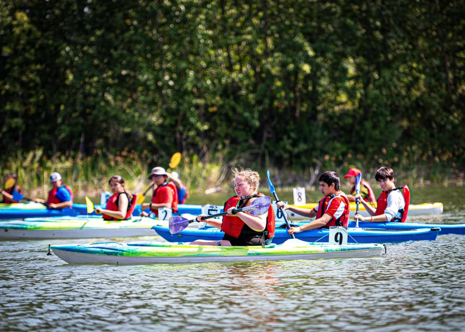 Kayakers wearing life jackets paddle numbered boats on calm water near a forested shoreline.