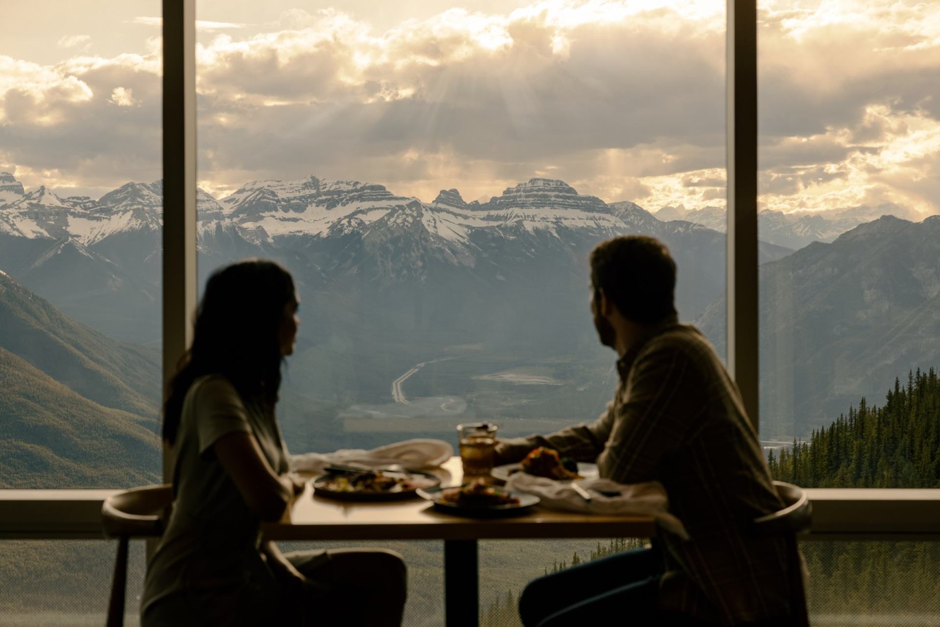 Silhouetted diners sit at a window table overlooking snow-capped mountain range.